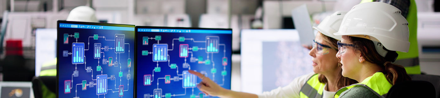 engineers in hard hats in production facility discussing a program on computer monitors